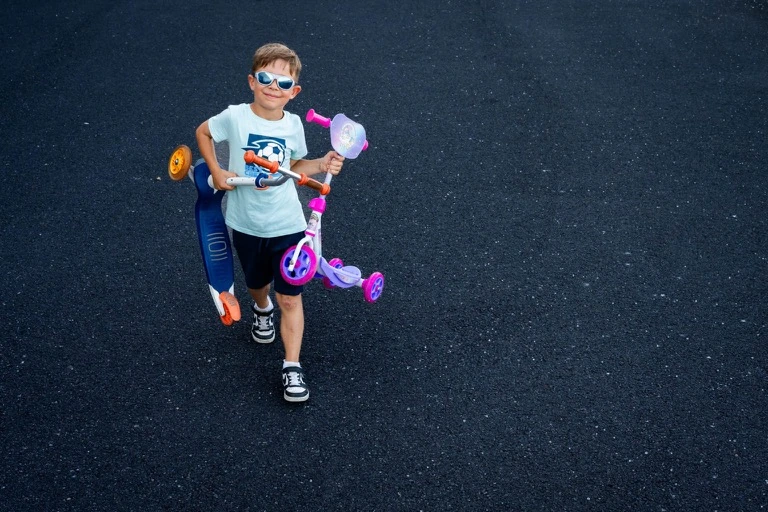 Un garçon en lunettes de soleil porte une trottinette violette sur l’épaule