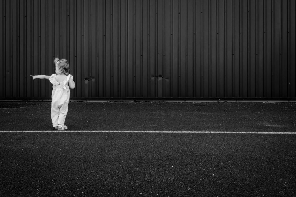 Une petite fille tend les bras devant un grand mur, en noir et blanc
