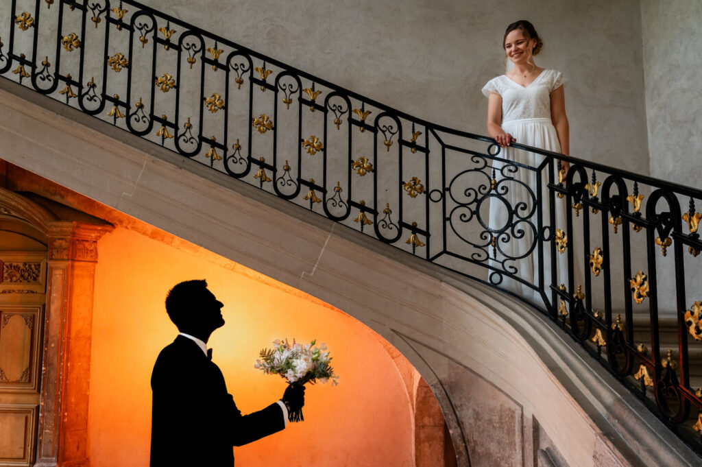 Une mariée en haut d’un escalier face au marié tenant son bouquet