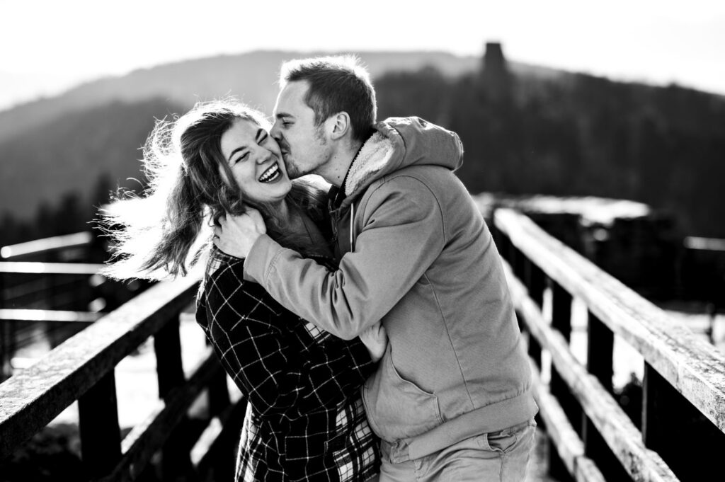 Un couple sourit et s’embrasse sur une passerelle en bois, photographié en noir et blanc.