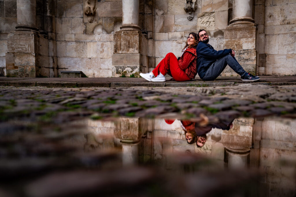 Un couple assis devant une façade en pierre se reflète dans une flaque au premier plan.