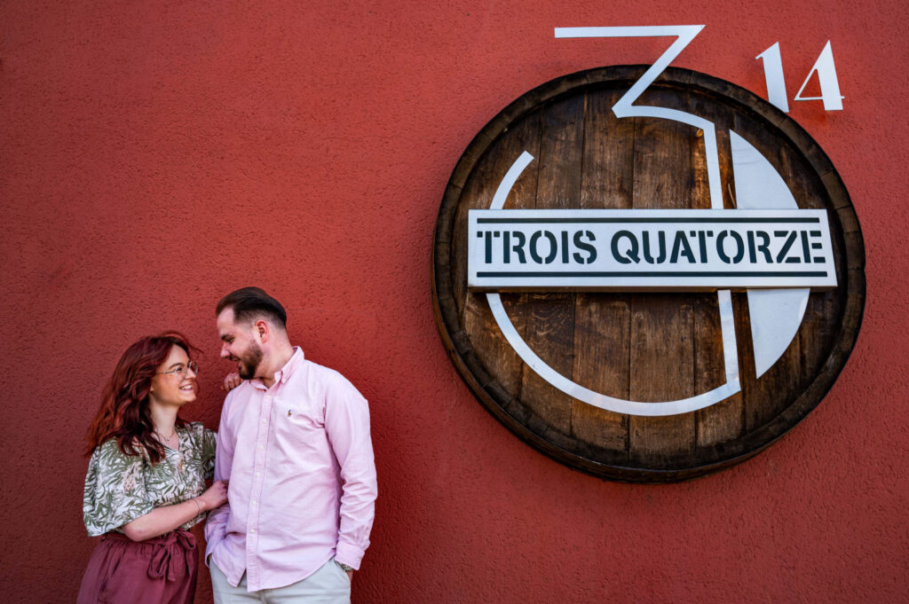 Un couple échange un regard et un sourire devant une façade rouge et une enseigne ronde.