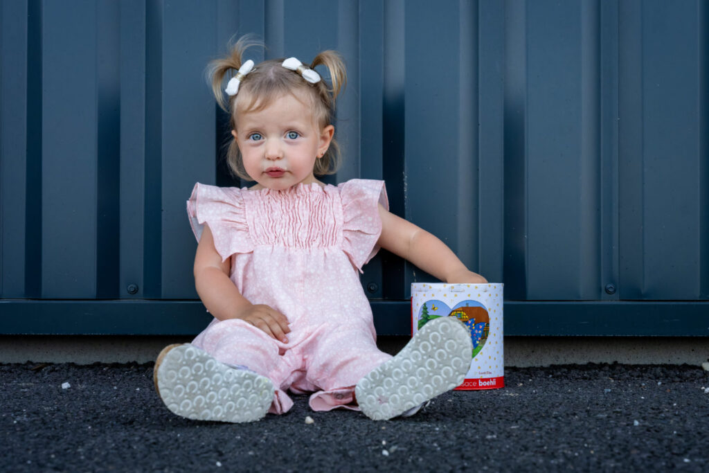 Une petite fille en salopette rose est assise devant un mur bleu