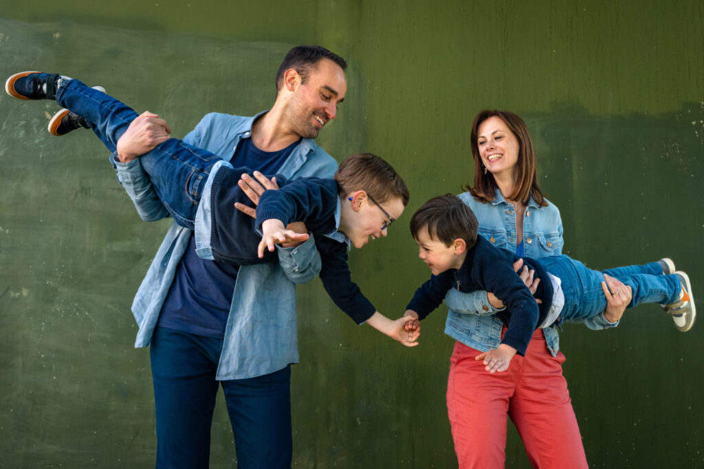 Des parents portent leurs deux enfants à l’horizontale devant un mur vert