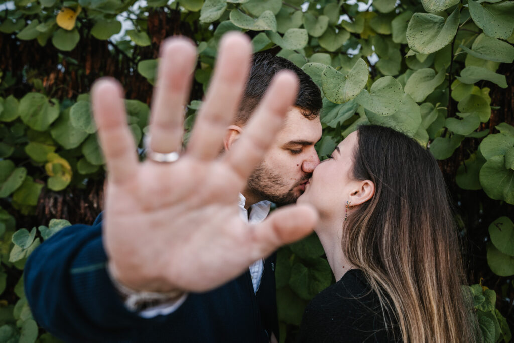 Un couple s’embrasse devant du feuillage en tendant une main vers l’objectif pour montrer une bague.